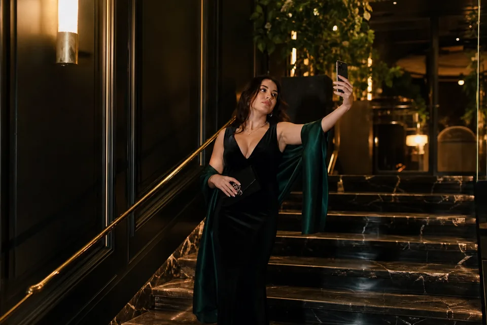 Adult woman in a long black velvet evening dress taking a selfie on a dark marble staircase at night