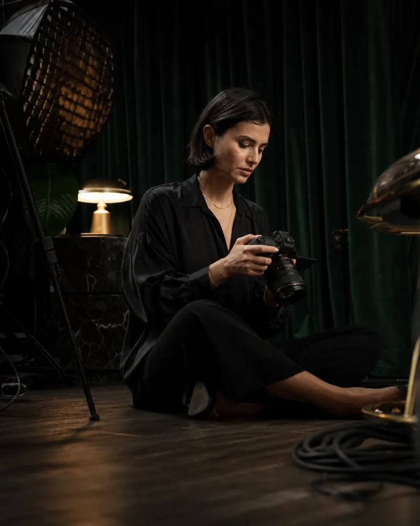 Adult woman in a black silk shirt reviewing video clips on a camera screen in her home studio with velvet curtains